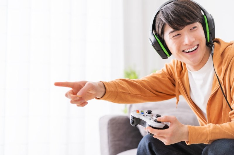 ヘッドホンをしてゲームコントローラーを持った男性が笑顔で指をさす様子